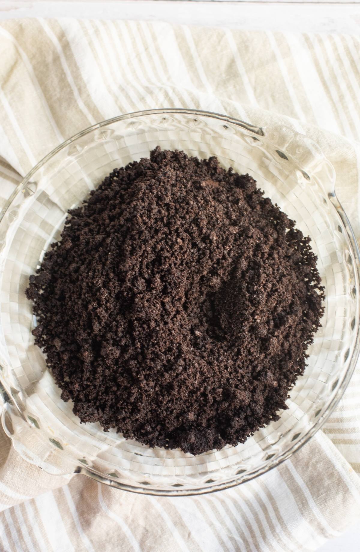 A glass bowl filled with crushed chocolate cookie crumbs, perfect for making a chocolate graham crust for peanut butter cream pie, sits on a striped cloth.