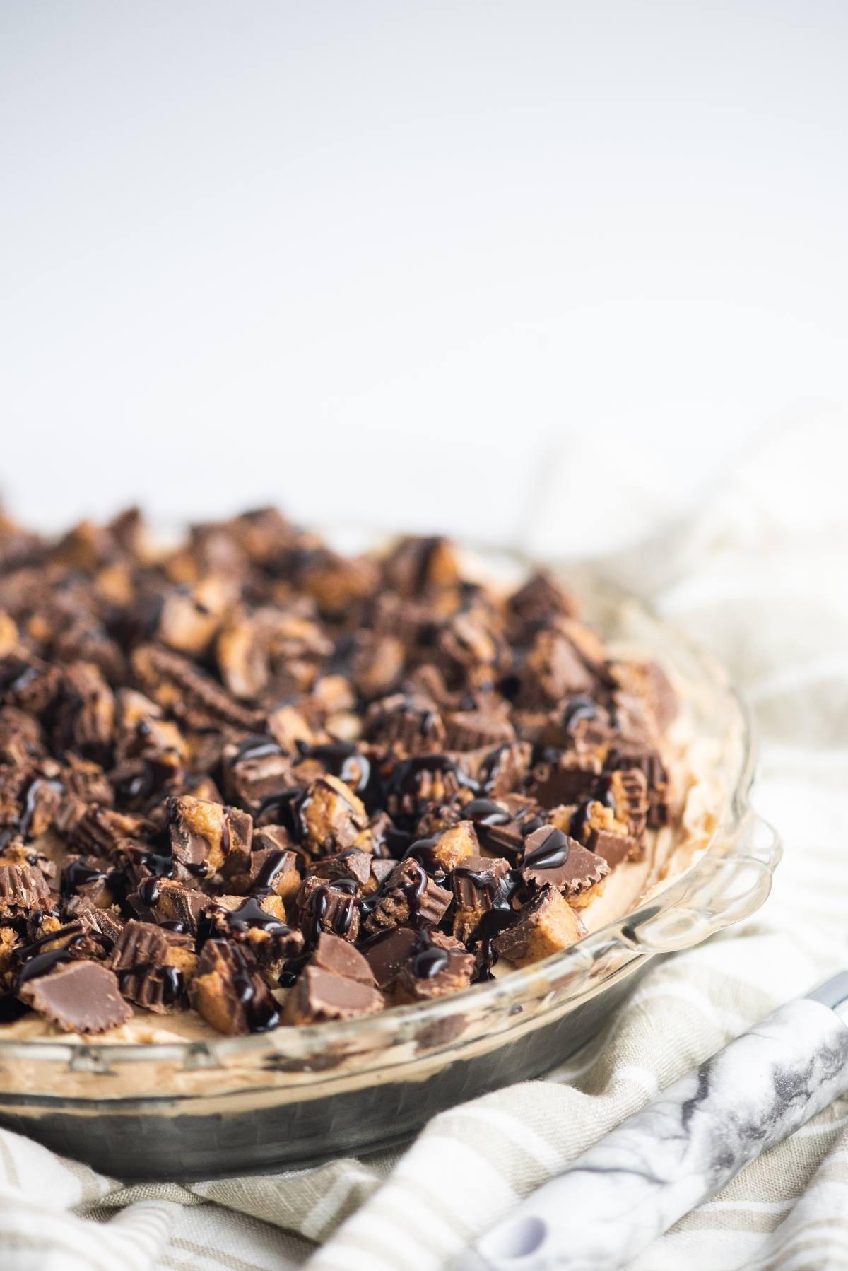 A peanut butter cream pie in a glass dish, topped with chopped peanut butter cups and chocolate drizzle, all on a chocolate graham crust.