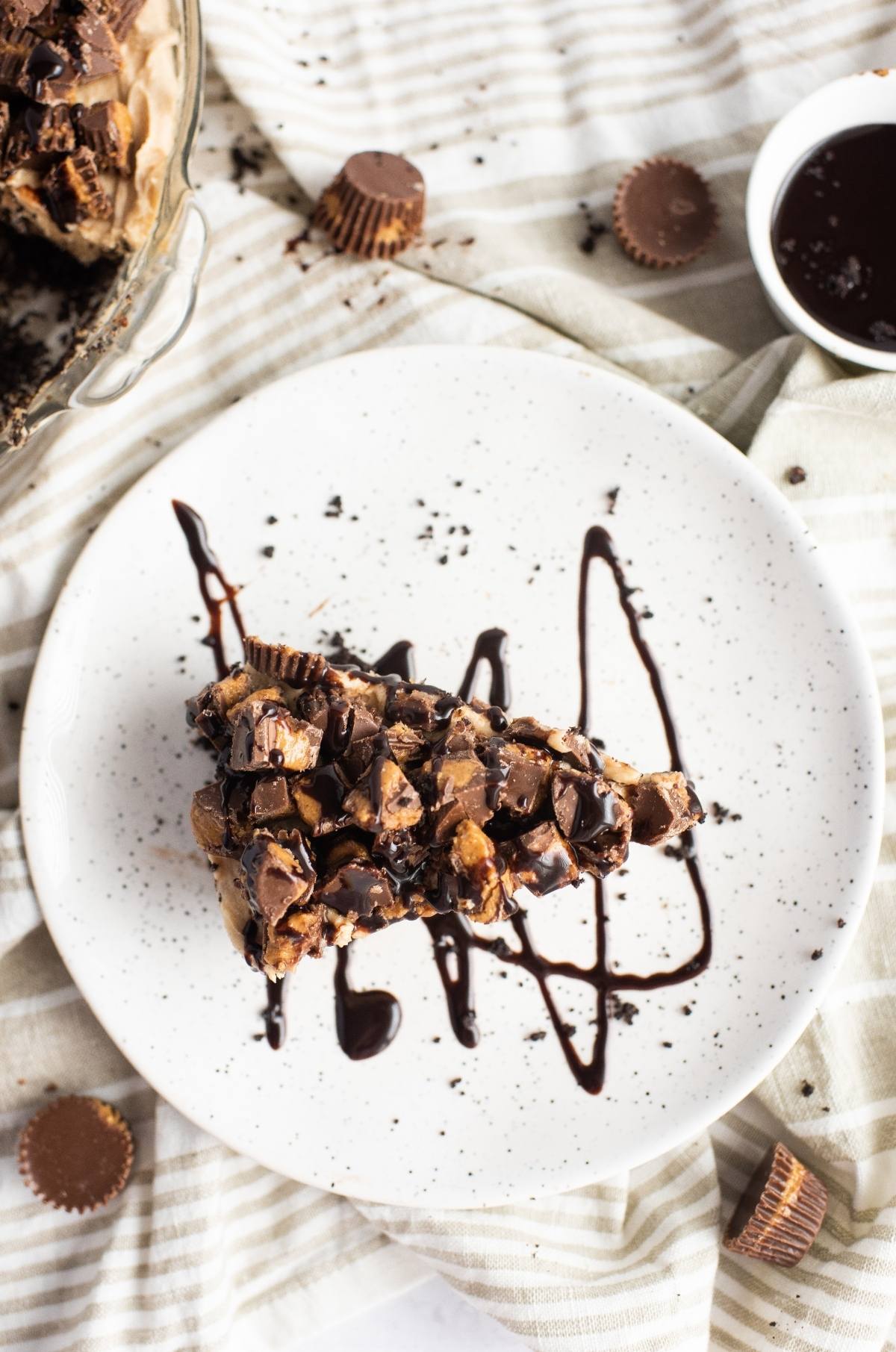 A slice of rich chocolate peanut butter pie with chocolate drizzle and peanut butter cups is served on a white plate.