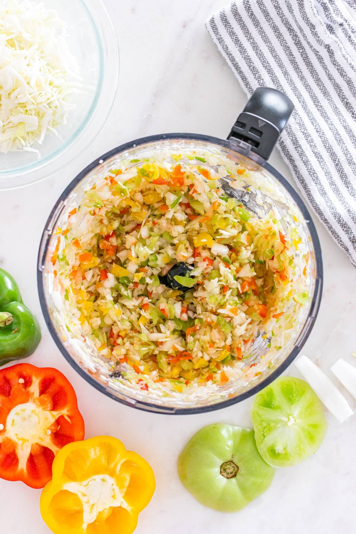 Chopped vegetables in a food processor, with bell peppers and a green tomato on a white countertop.