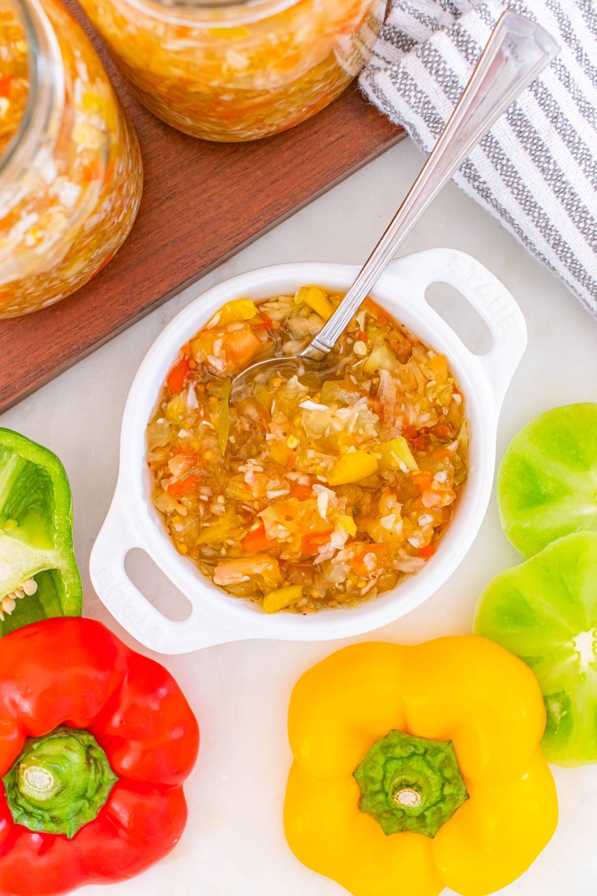 A small white dish of homemade relish with a spoon, surrounded by red, yellow, and green bell peppers.