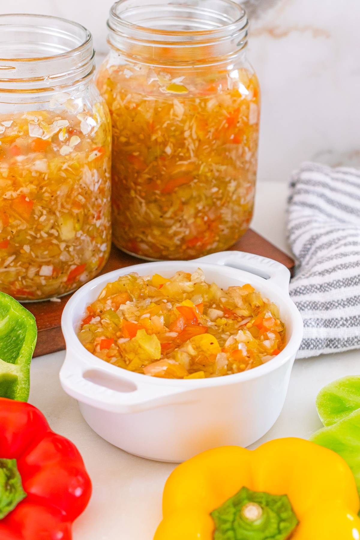 Chopped vegetable relish in a white bowl, with jars of relish and colorful bell peppers around it.