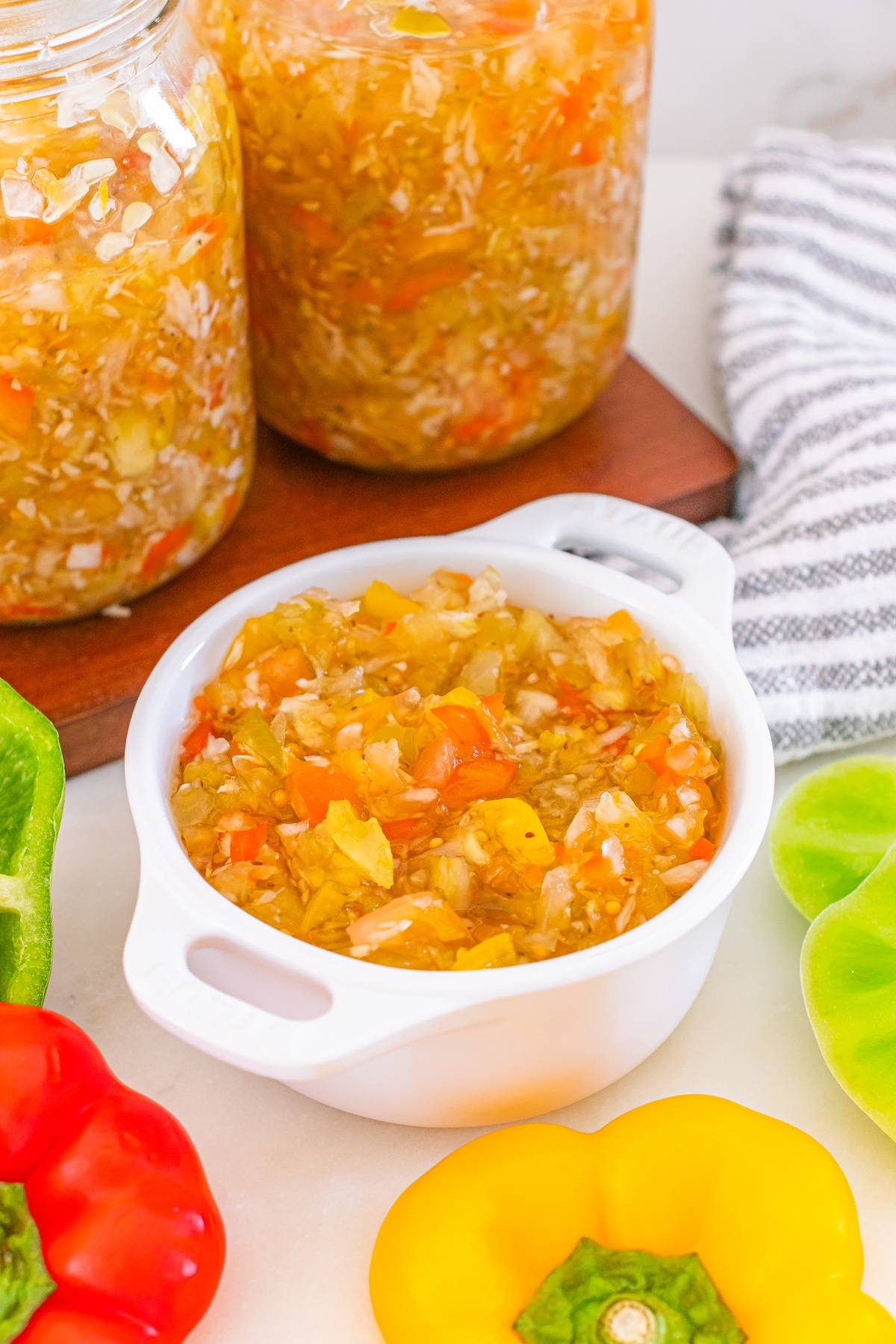 A white bowl of homemade relish with chopped peppers and onions, surrounded by fresh bell peppers.
