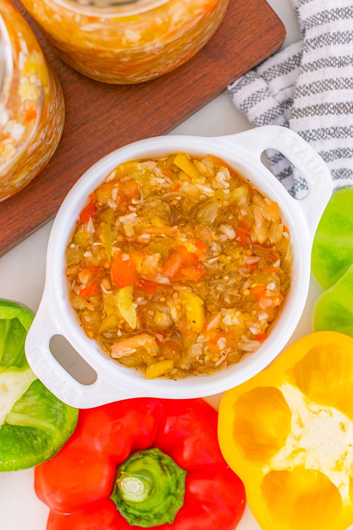 A white bowl of chopped pickled peppers and onions, surrounded by fresh green, red, and yellow bell peppers.