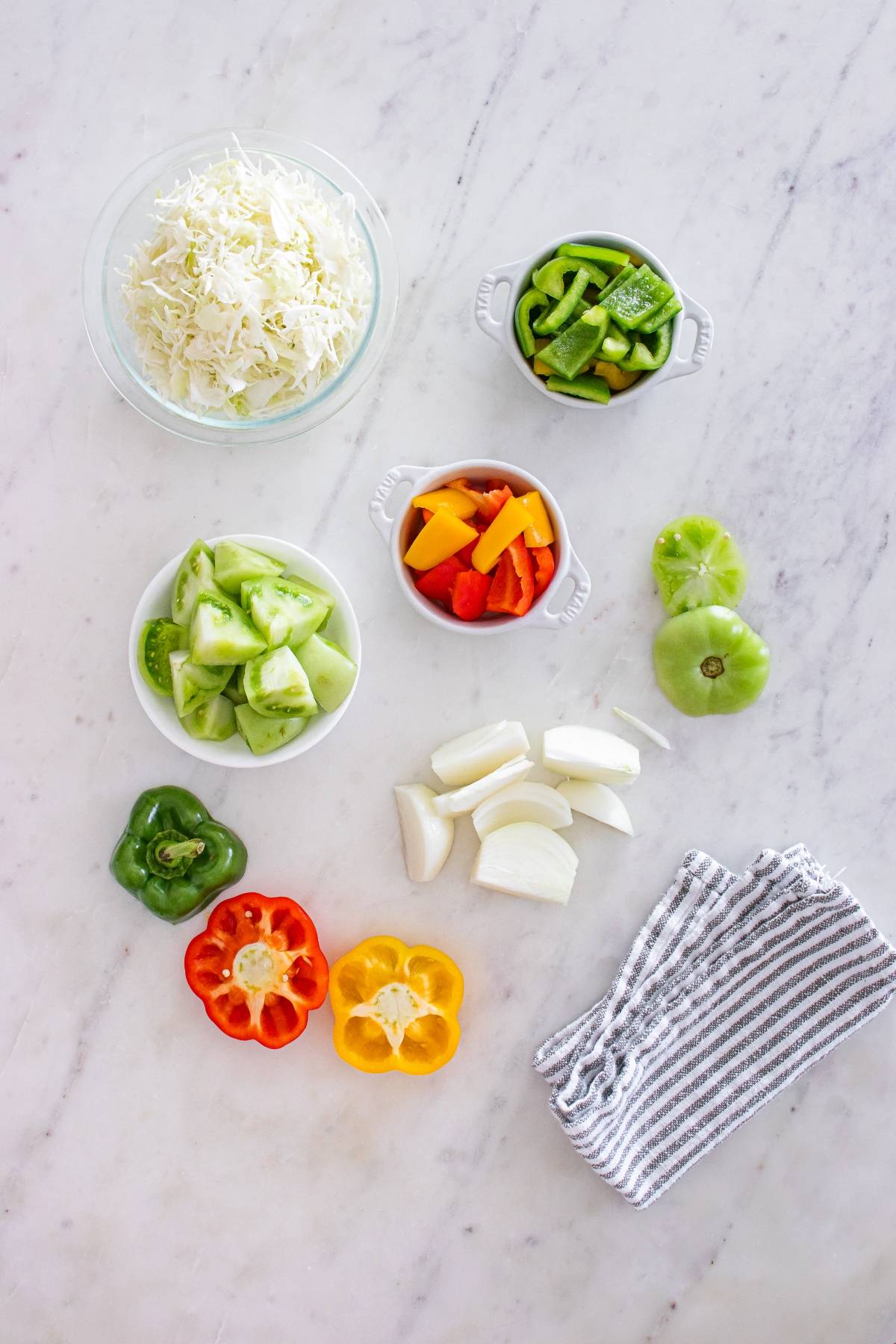 Chopped cabbage, tomatoes, onions, and sliced bell peppers arranged on a marble countertop with a striped towel.