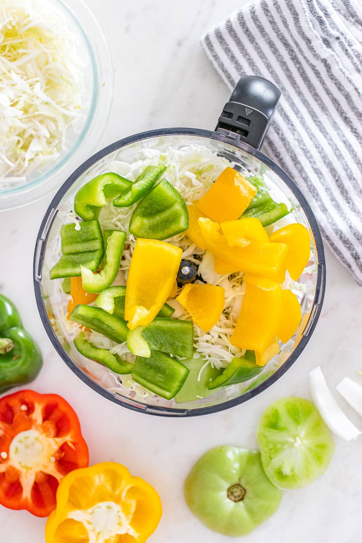 Chopped green and yellow bell peppers in a food processor with shredded cabbage, surrounded by fresh veggies.