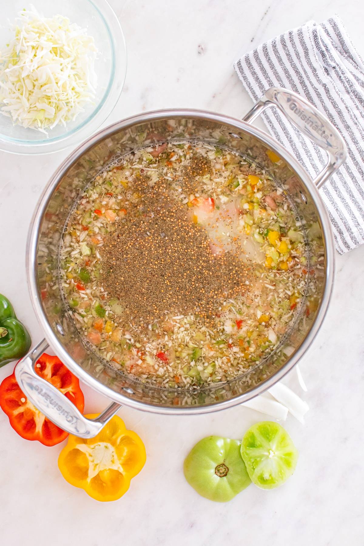 Overhead view of a pot with chopped vegetables and seasonings, surrounded by sliced peppers and a dish towel.
