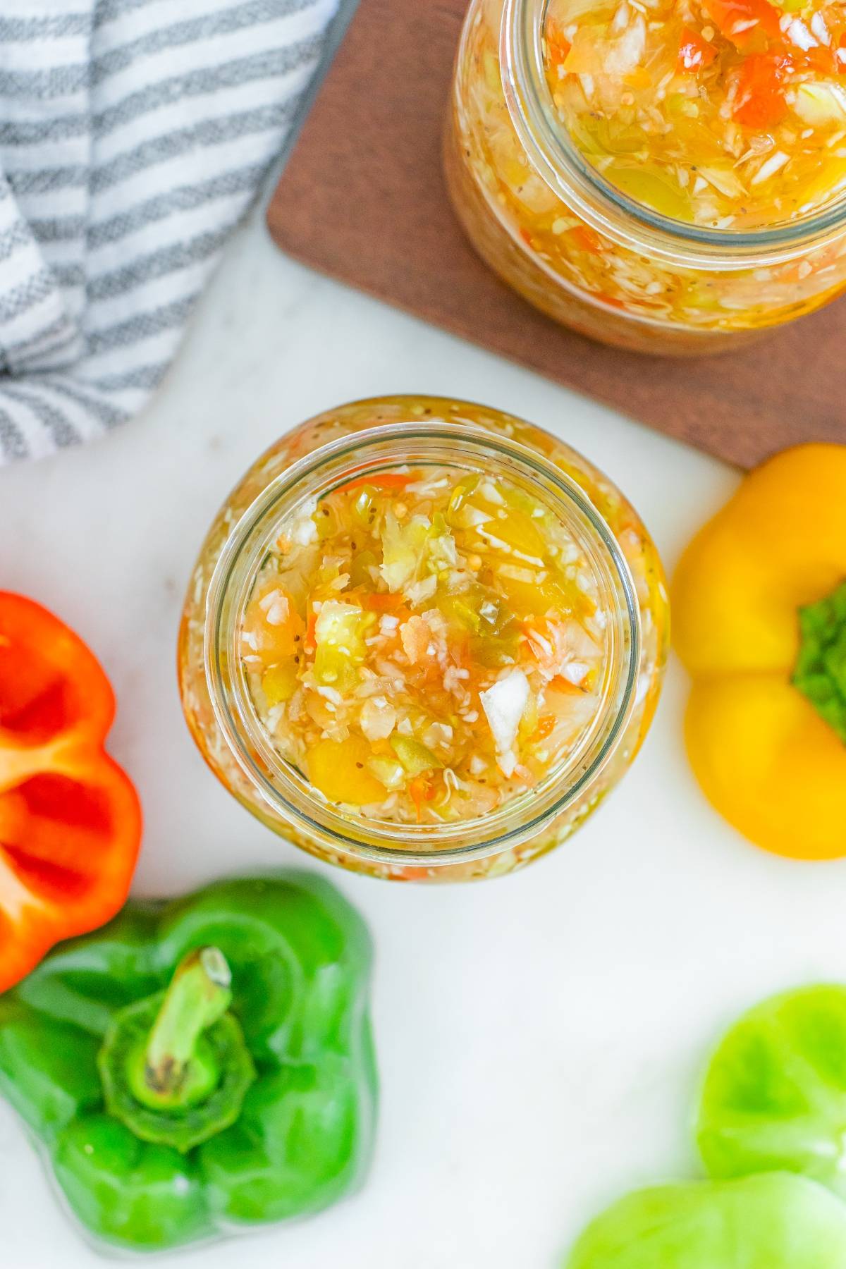 Top view of a jar filled with chopped peppers and onions, surrounded by fresh bell peppers on a white surface.