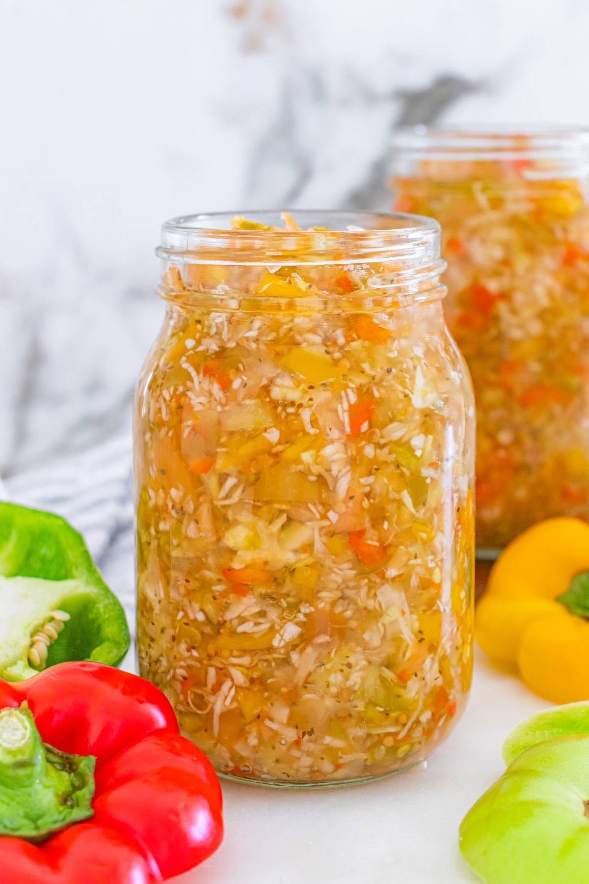 A jar of homemade relish with chopped peppers and vegetables, surrounded by fresh bell peppers.
