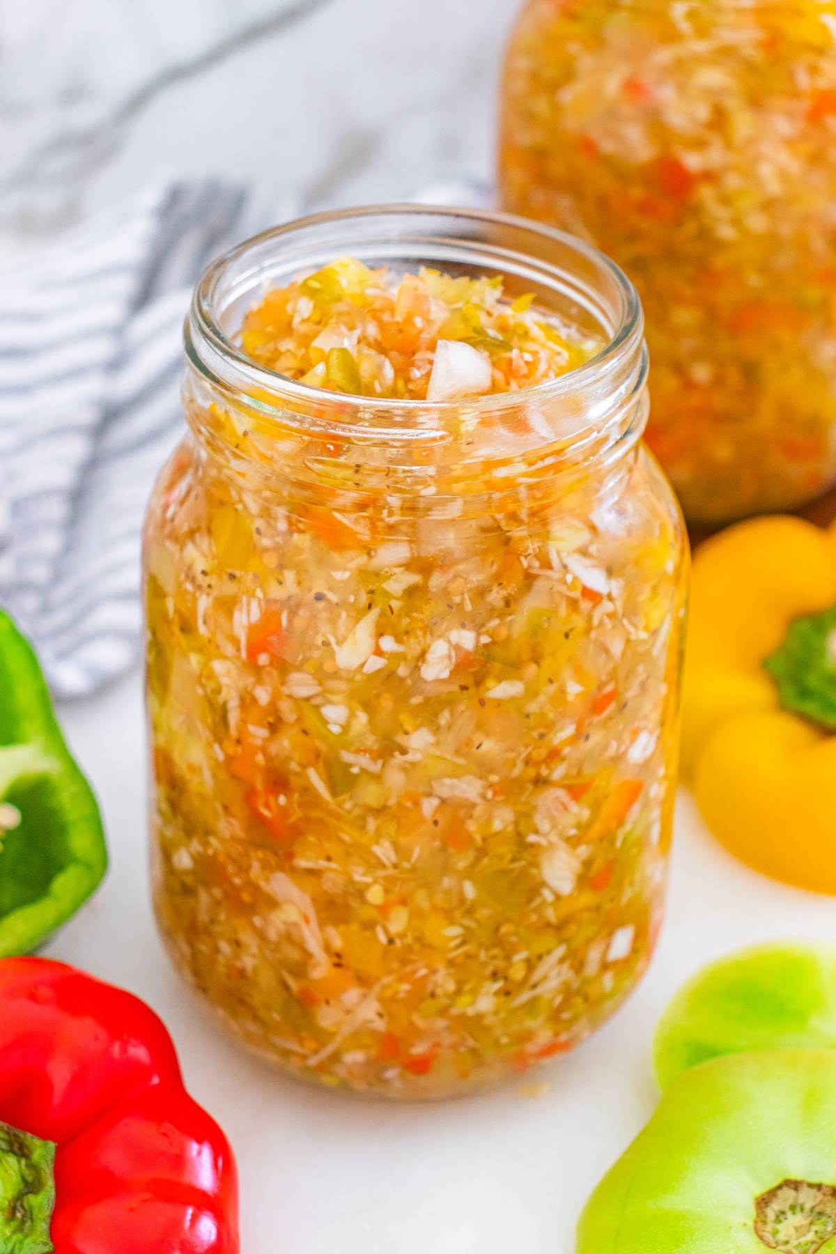 A glass jar filled with colorful homemade relish, surrounded by fresh bell peppers and a napkin.