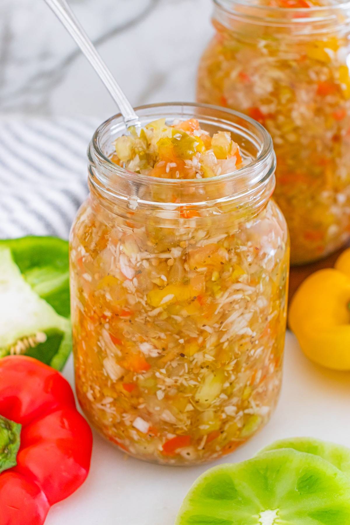 A glass jar filled with chopped vegetable relish, surrounded by colorful bell peppers.