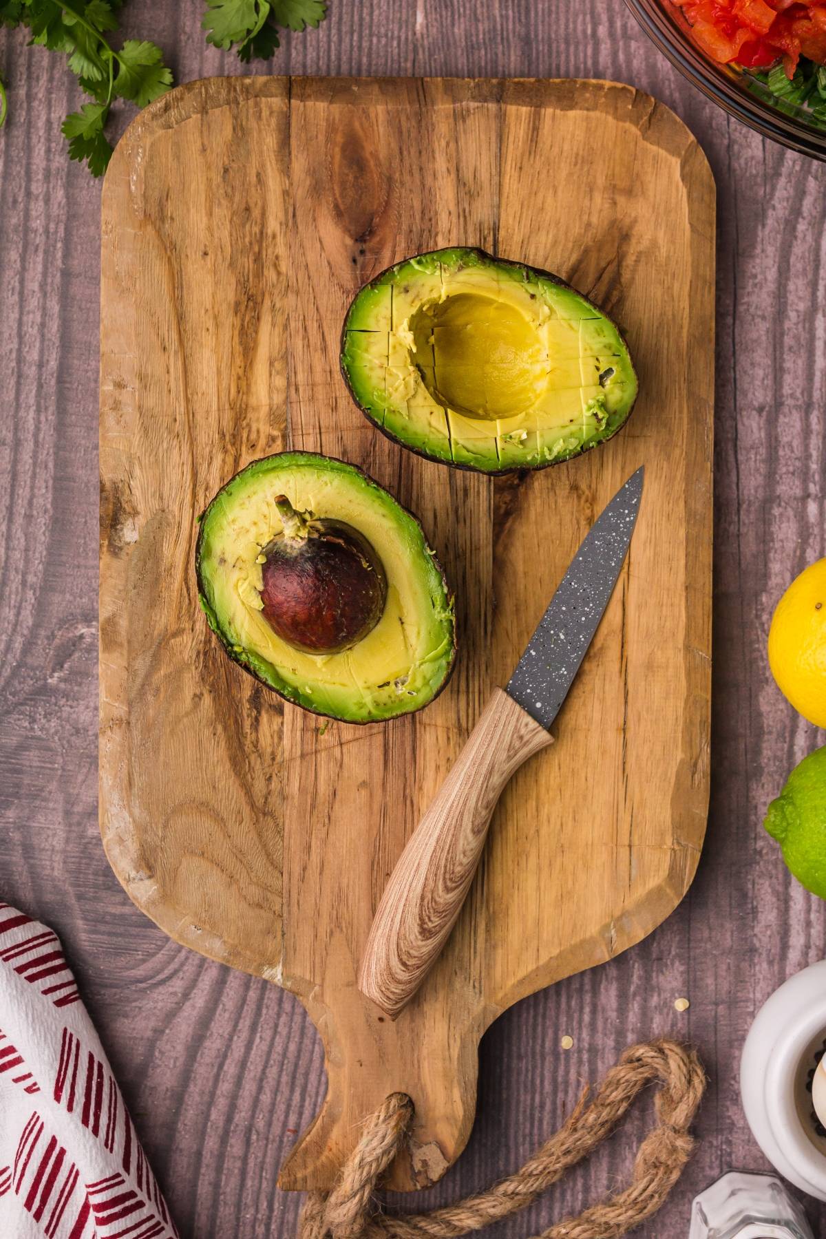 A halved avocado and a knife on a wooden cutting board, surrounded by fruits and herbs—perfect ingredients for an easy guacamole recipe.