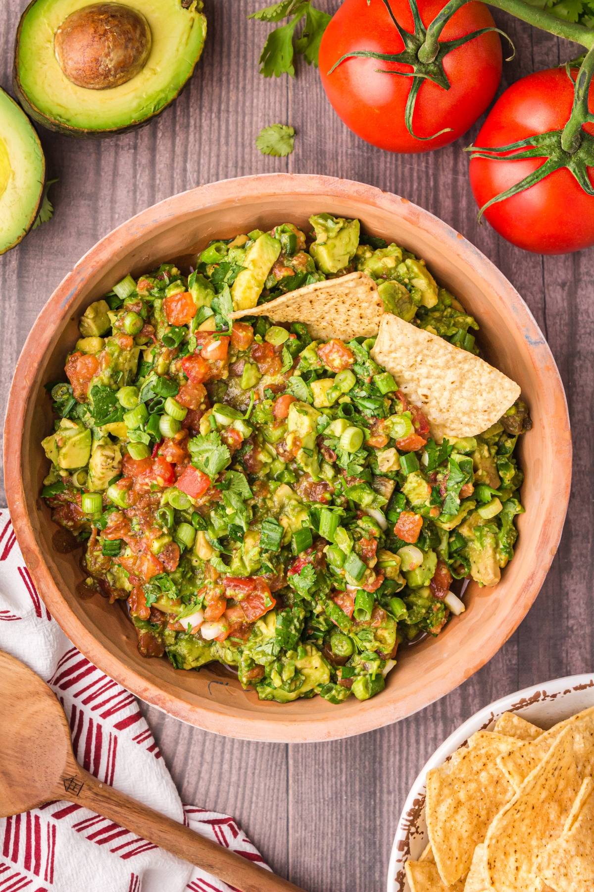 Bowl of chunky guacamole made from an easy guacamole recipe, served with tortilla chips and surrounded by tomatoes, avocado, and chips on a wooden table.
