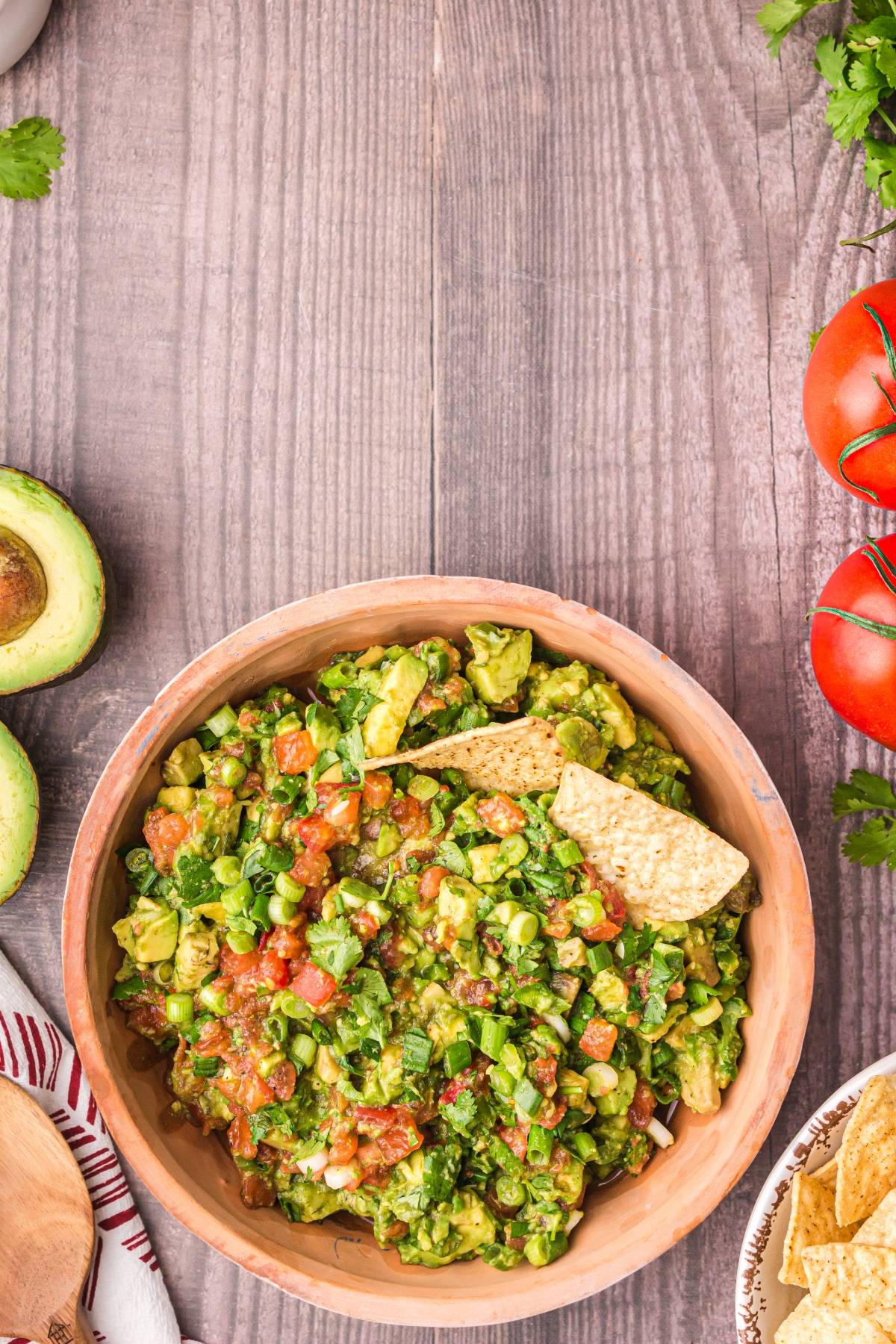 A bowl of chunky guacamole with tortilla chips, surrounded by fresh avocado, tomatoes, and cilantro on a wooden table.