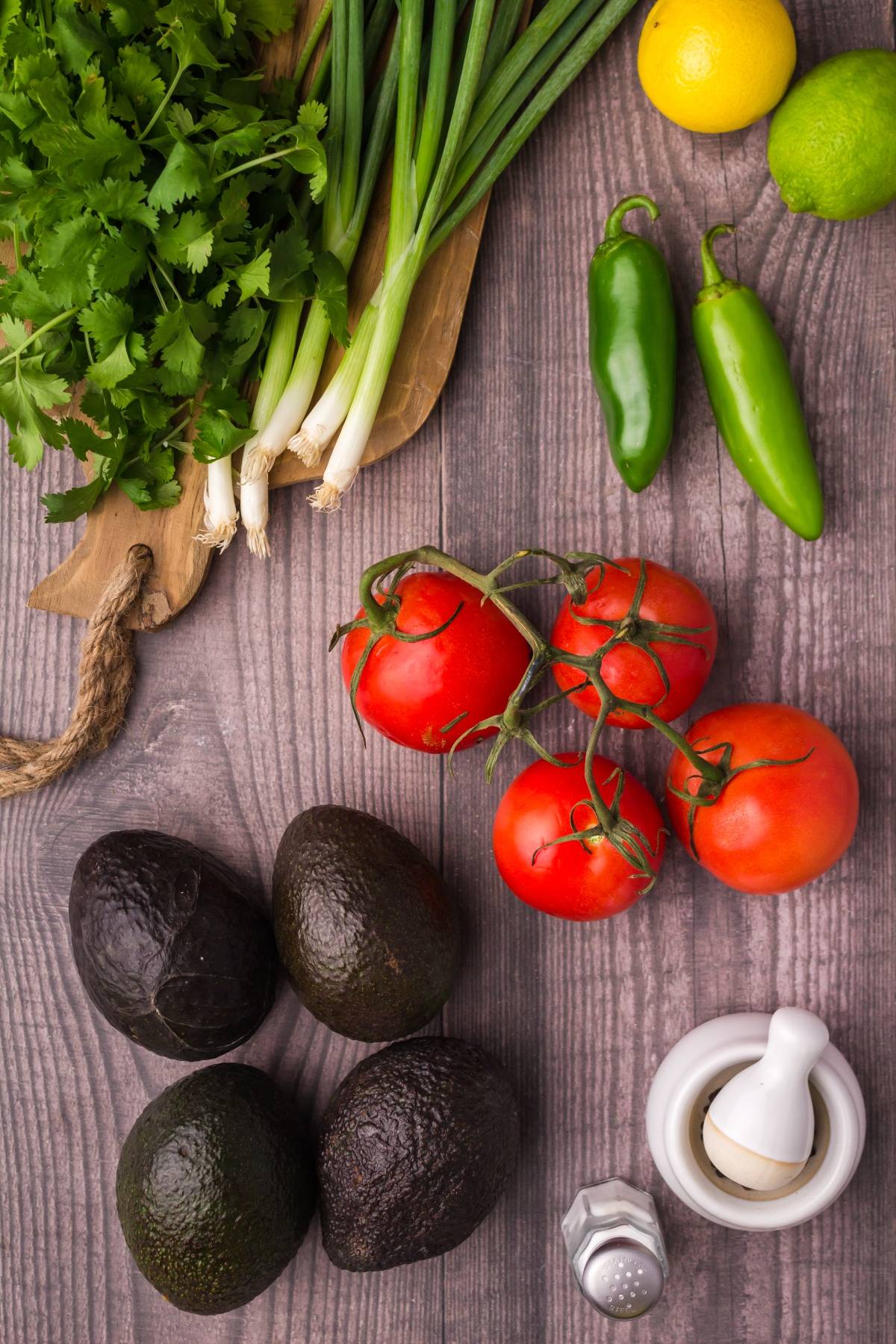 Fresh cilantro, scallions, tomatoes, avocados, green peppers, lemon, lime, salt, and mortar on a wooden table—perfect ingredients for delicious homemade guacamole or a chunky guacamole dip.