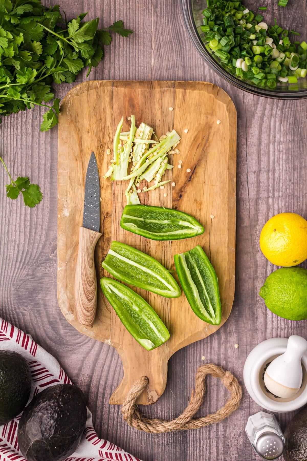 Sliced jalapeños and a knife on a wooden cutting board, surrounded by fresh herbs, lemon, lime, and avocados—perfect ingredients for homemade guacamole.