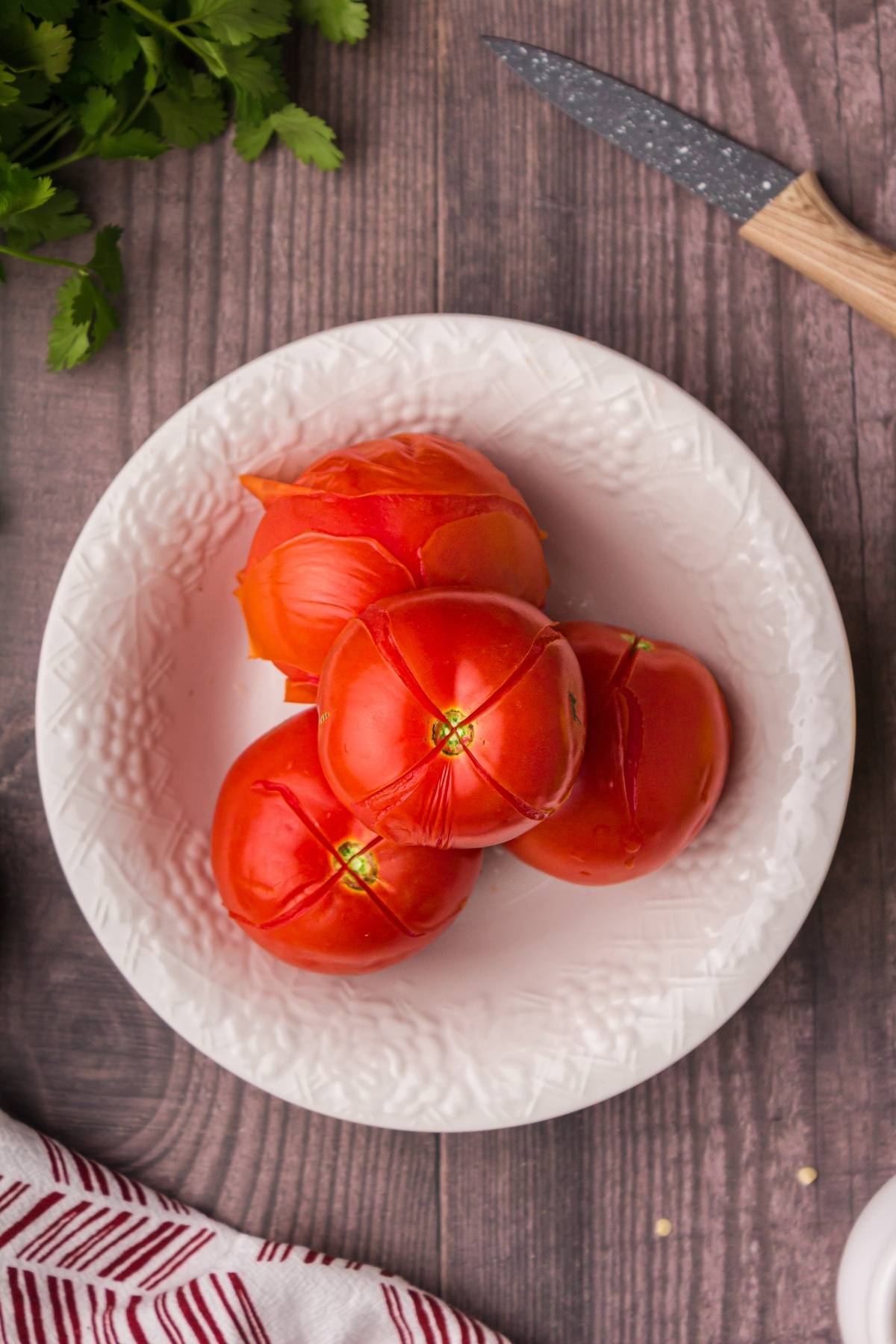 Four peeled tomatoes in a white bowl on a wooden table, with a knife and herbs nearby—perfect ingredients to start your homemade guacamole or add to your favorite easy guacamole recipe.