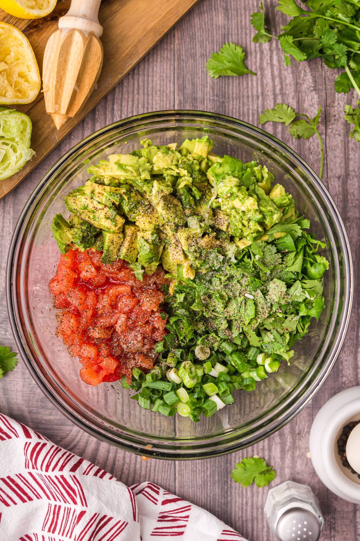 A glass bowl with chopped avocado, tomatoes, green onions, and cilantro on a wooden table—everything you need for homemade guacamole, ready to mix.