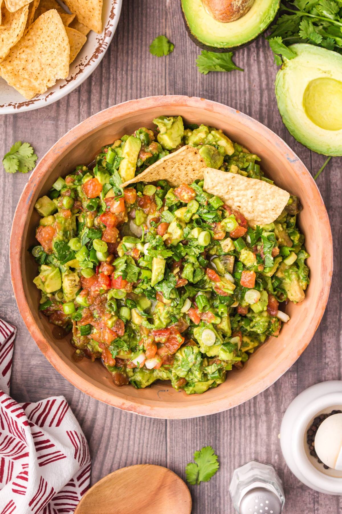A bowl of chunky guacamole with tortilla chips, surrounded by avocado, cilantro, and a bowl of chips on a wooden table.