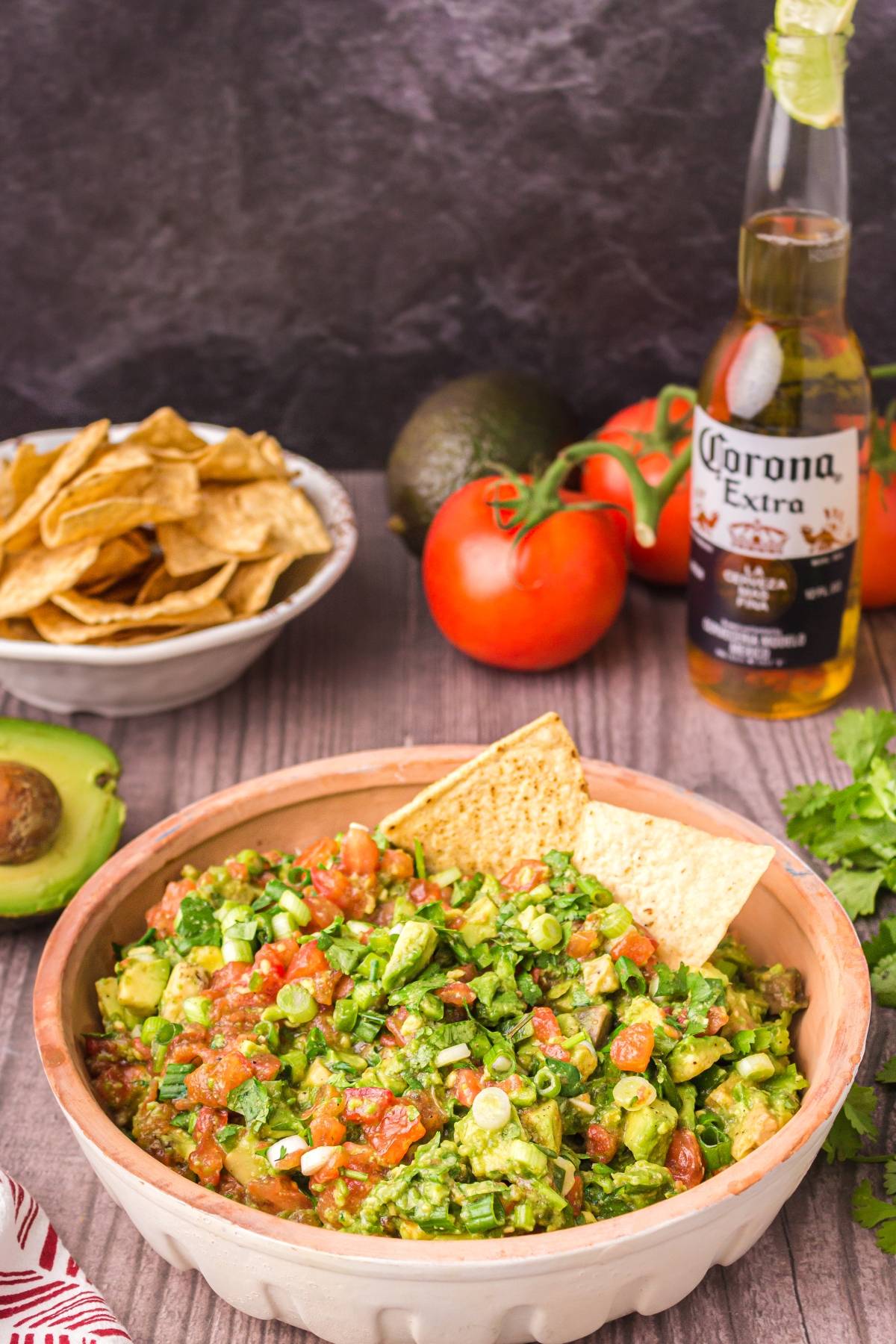 Bowl of chunky guacamole with tortilla chips, fresh tomatoes, avocado, cilantro, chips, and a Corona beer in the background—an easy guacamole recipe perfect for snacking.