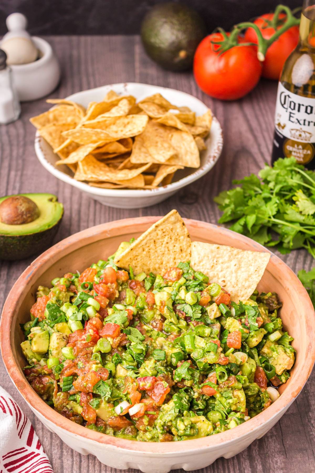 Bowl of chunky guacamole made with our easy guacamole recipe, served with tortilla chips and surrounded by tomatoes, avocado, cilantro, and a bottle of beer.
