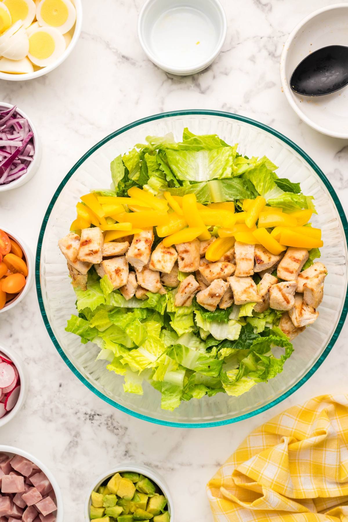 A filling dinner salad with lettuce, grilled chicken, and yellow peppers is served in a bowl, surrounded by sliced veggies and perfect with homemade ranch dressing on a marble surface.