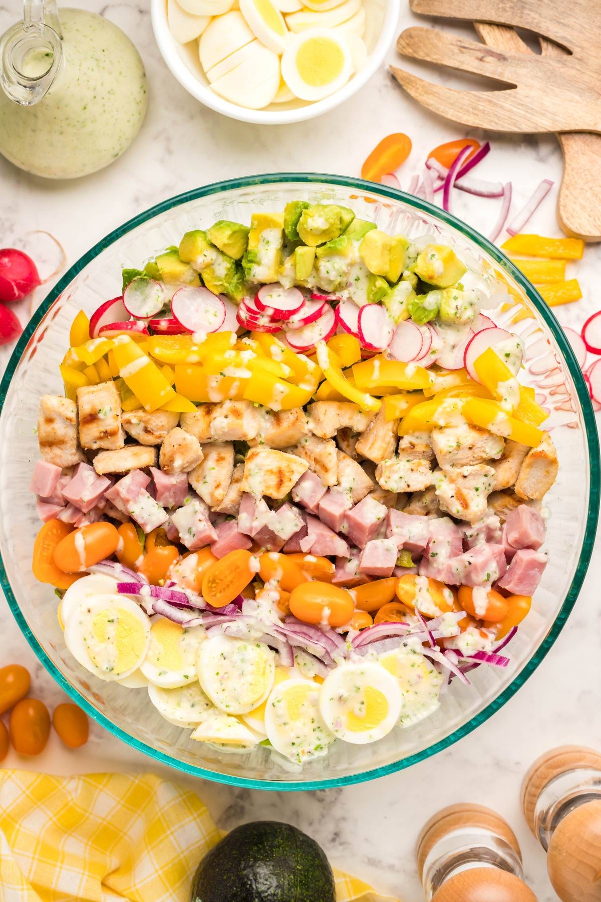 A glass bowl of Cobb salad with rows of ham, chicken, eggs, tomatoes, radishes, onions, and homemade ranch dressing.
