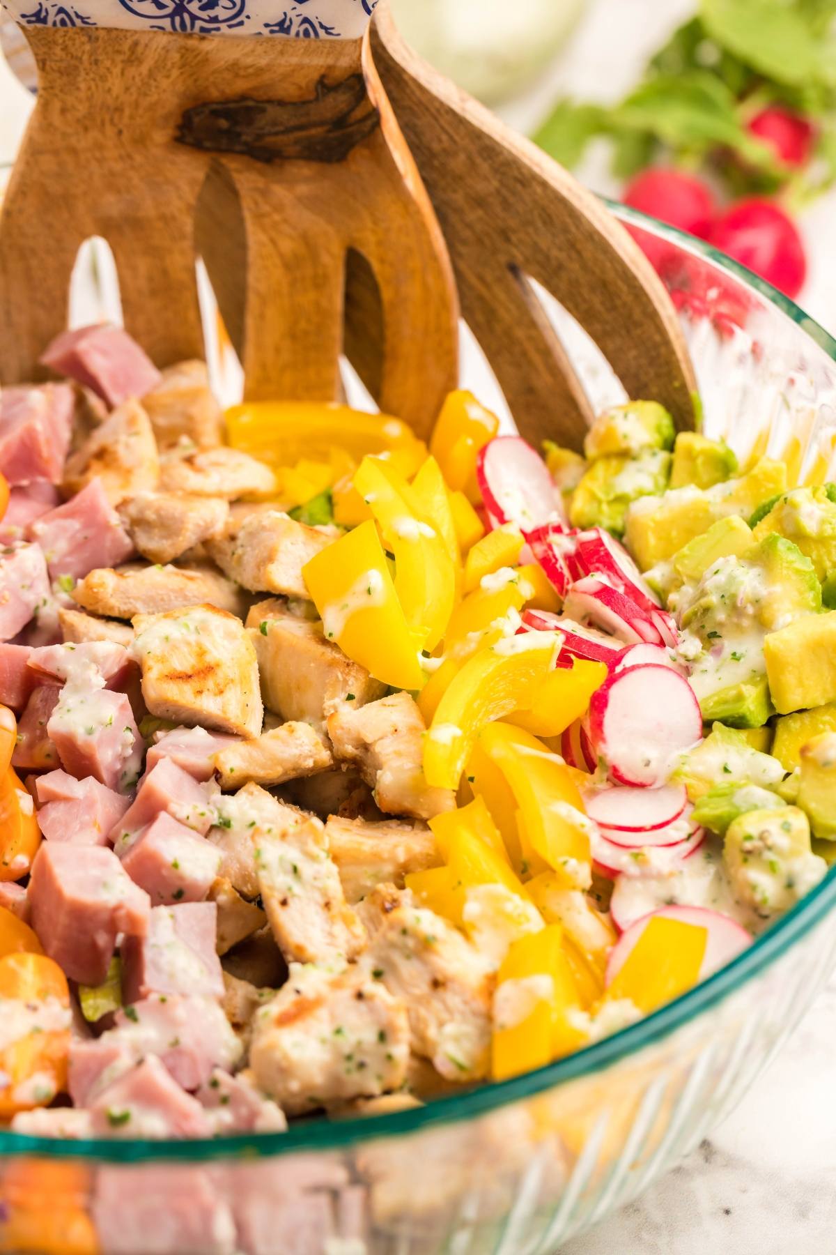 Chopped Cobb salad with ham, chicken, yellow peppers, radishes, and avocado in a glass bowl with wooden tongs—perfect as a recipe using leftover ham and paired with homemade ranch dressing.
