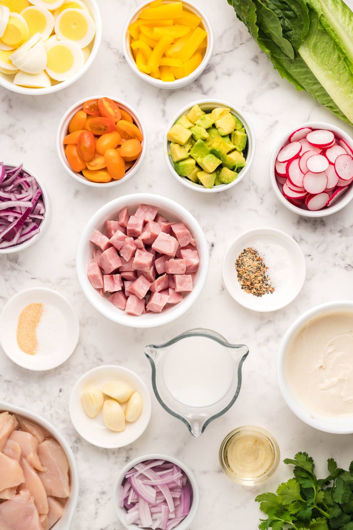 Bowls of chopped ham, vegetables, eggs, and homemade ranch dressing ingredients sit on a marble surface—perfect for assembling a classic Cobb salad.