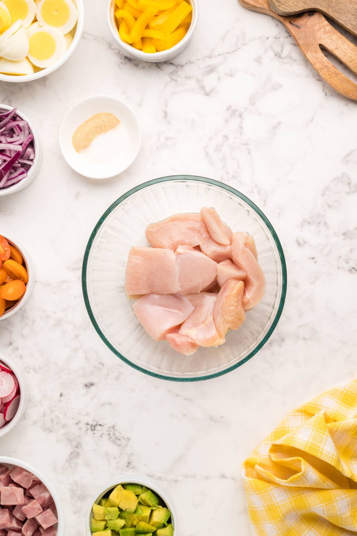 Glass bowl of raw chicken pieces surrounded by bowls of vegetables and seasonings on a marble countertop, perfect for preparing a hearty filling dinner salad or classic Cobb salad.