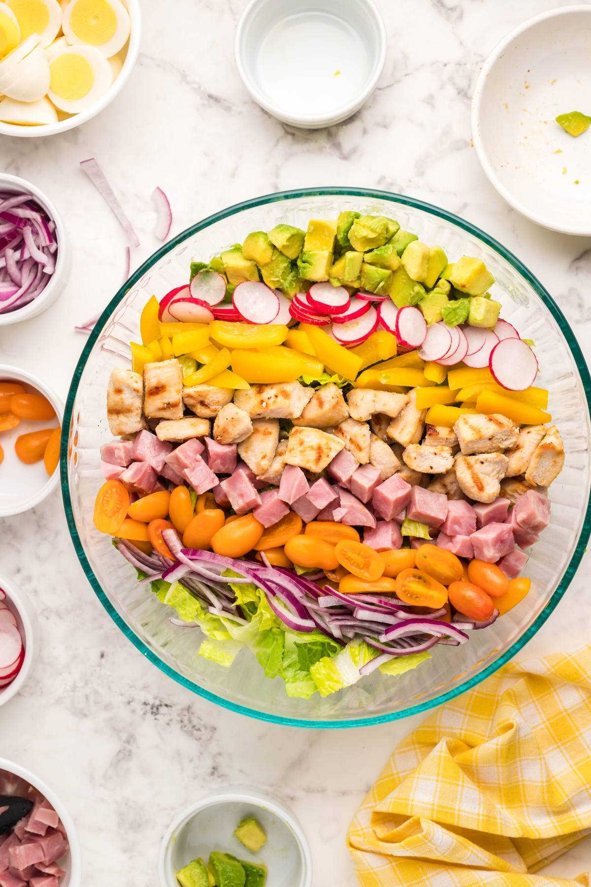 A glass bowl with layered salad ingredients: lettuce, ham, chicken, tomatoes, peppers, avocado, and radishes—perfect as a recipe using leftover ham and topped with homemade ranch dressing.