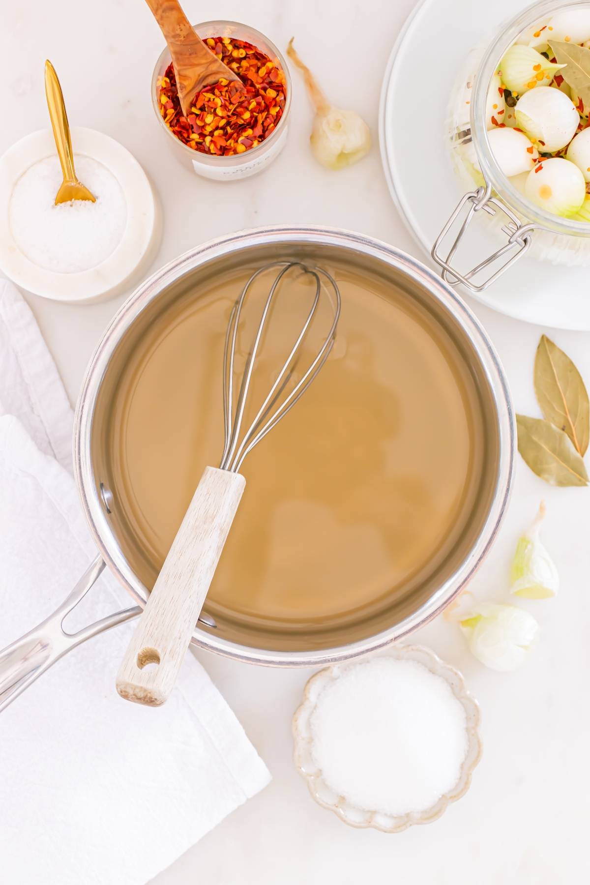 A saucepan of clear liquid with a whisk, surrounded by salt, spices, and a jar of pickling onions.