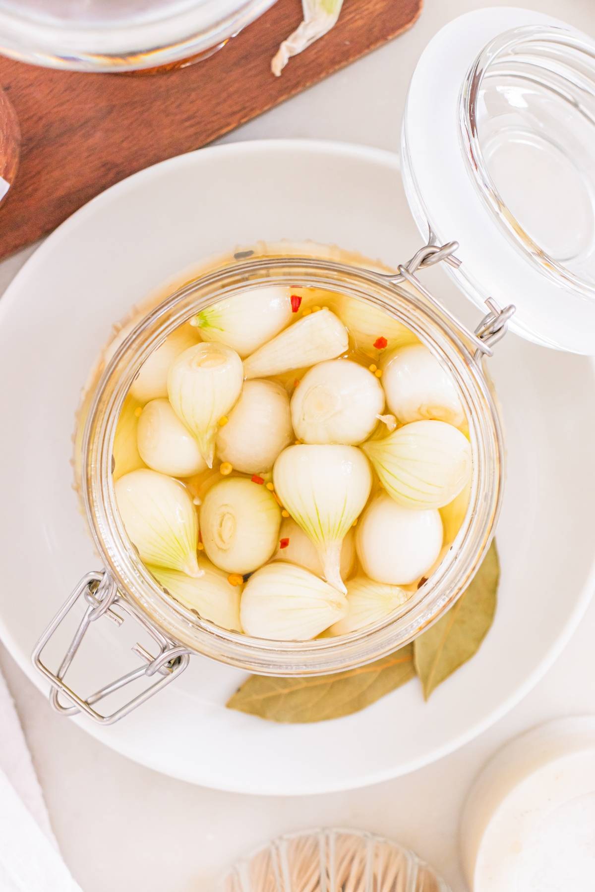 A glass jar filled with pickled pearl onions in brine, with a bay leaf and red pepper flakes visible.