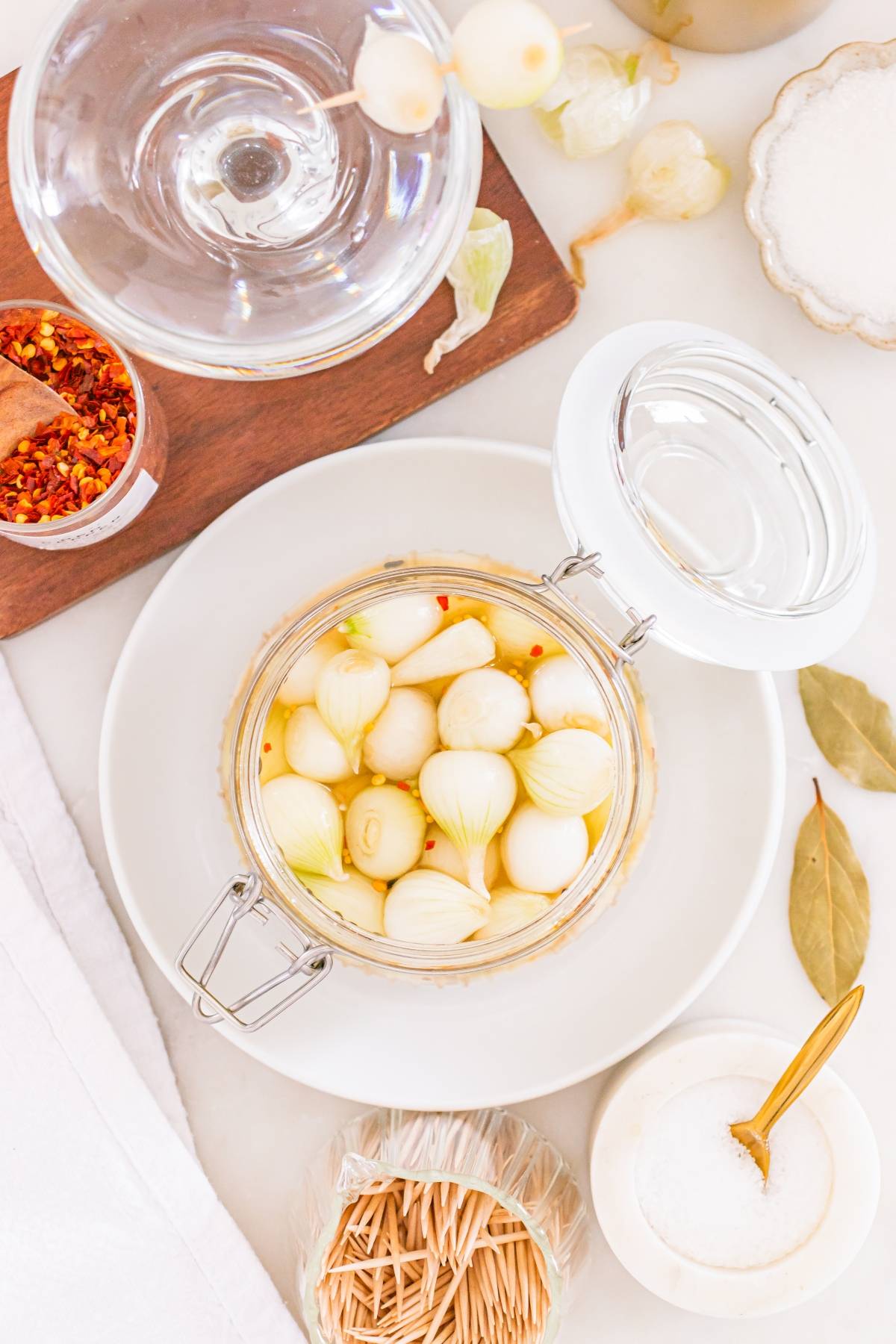 A jar of pickled pearl onions on a plate, surrounded by spices, bay leaves, and a glass of water.