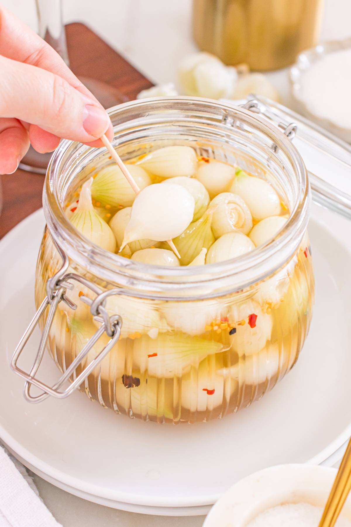 A hand uses a toothpick to pick up a pickled pearl onion from a glass jar filled with more onions in brine.