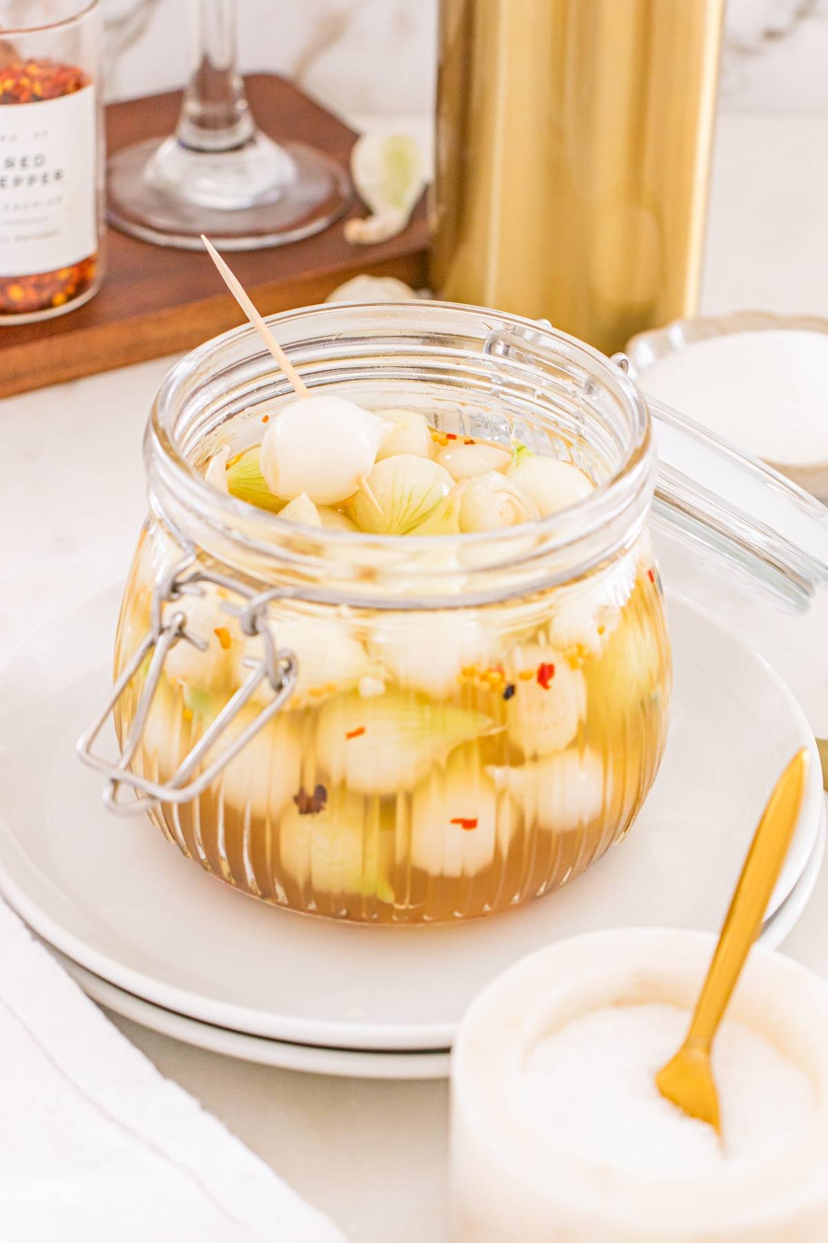 Glass jar filled with pickled onions in brine, with a toothpick, on a white plate beside a small bowl of salt.