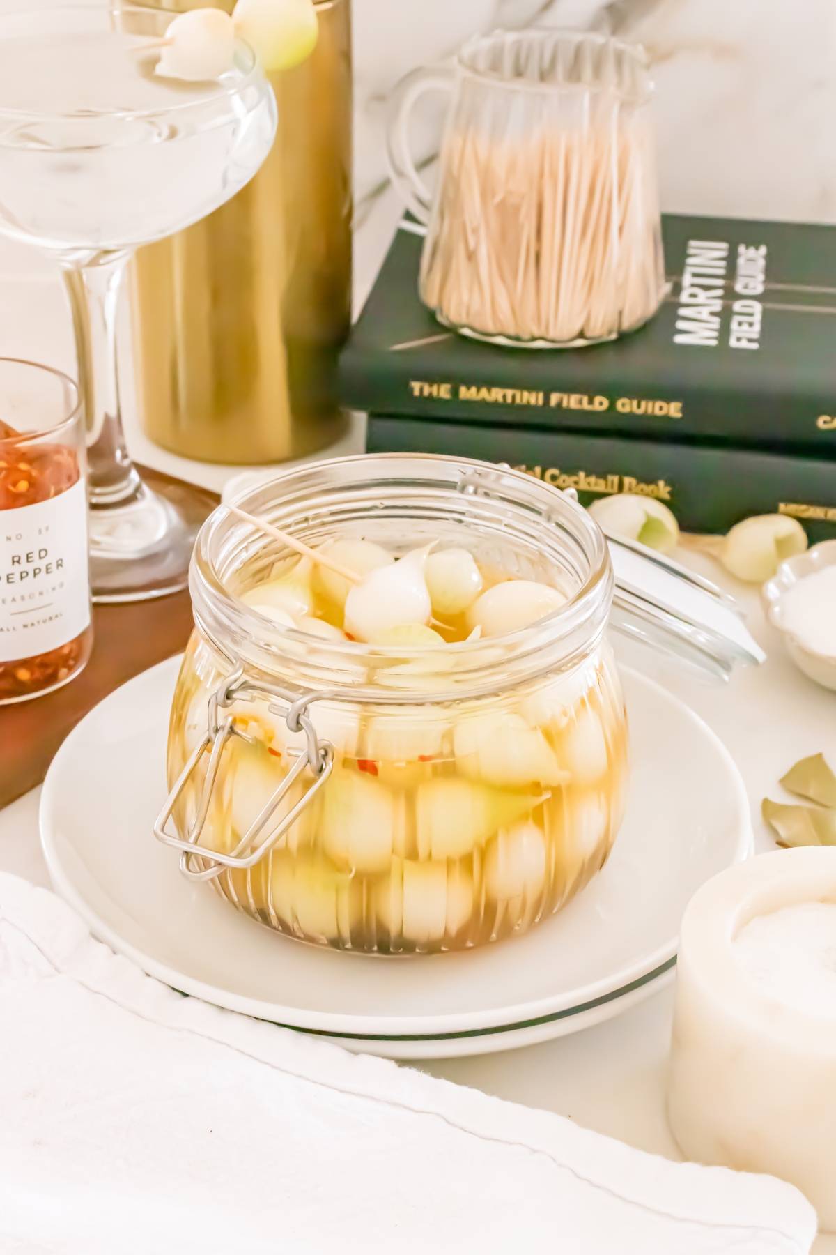 A jar of pickled cocktail onions on a plate, with martini books and barware in the background.