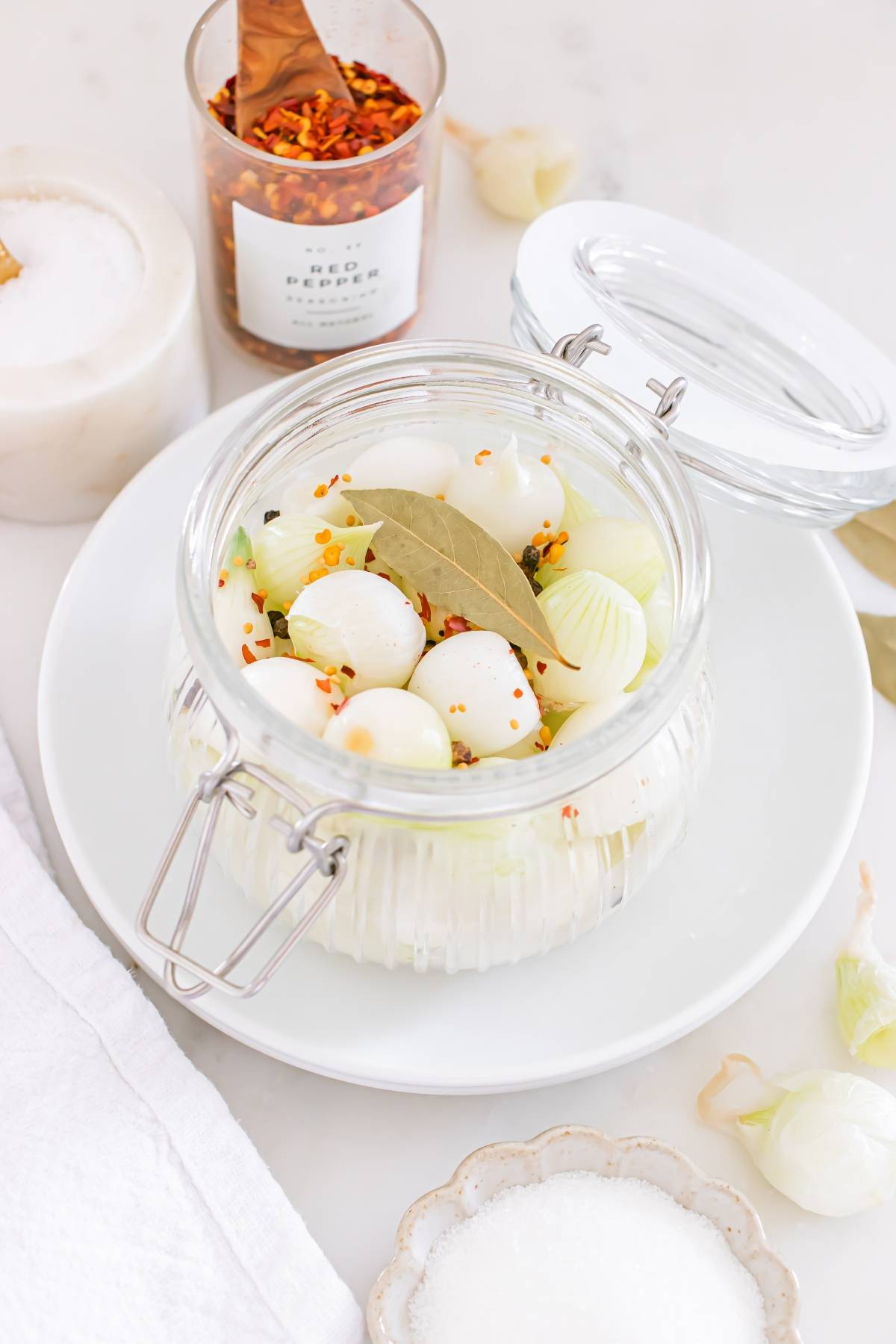 Glass jar with pearl onions, bay leaf, and chili flakes on a white plate, with seasoning jars nearby.