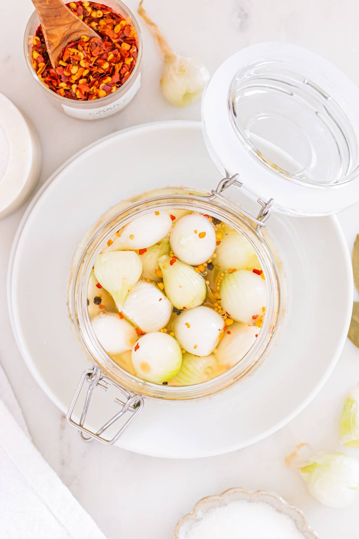 Glass jar of pickled pearl onions with chili flakes, surrounded by spices on a white surface.