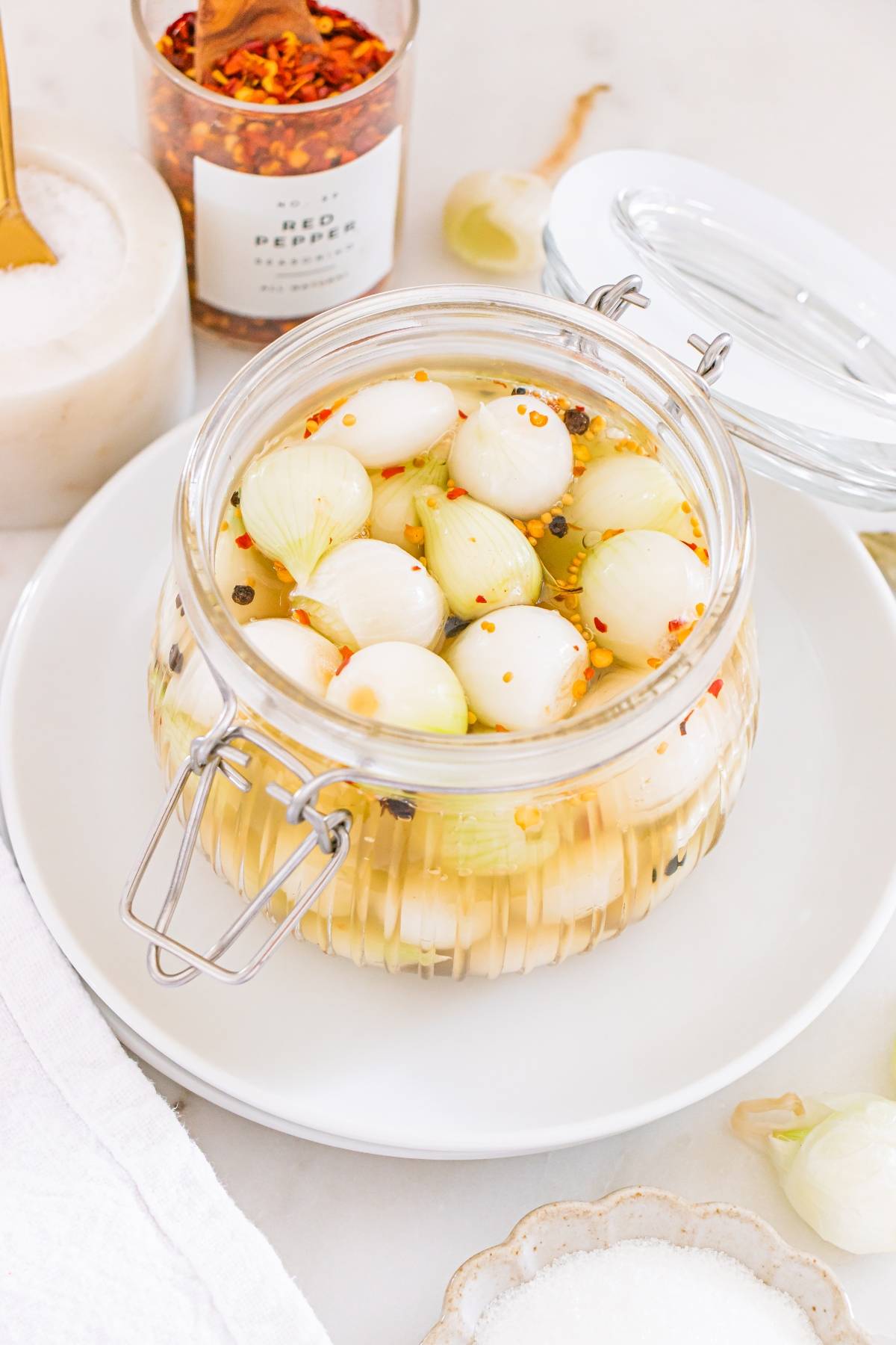A glass jar filled with pickled onions in brine, placed on a white plate beside a jar of red pepper flakes.