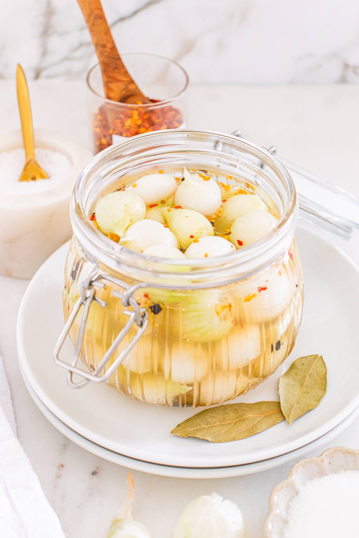 A jar of pickled pearl onions with bay leaves and spices, placed on a white plate on a marble surface.