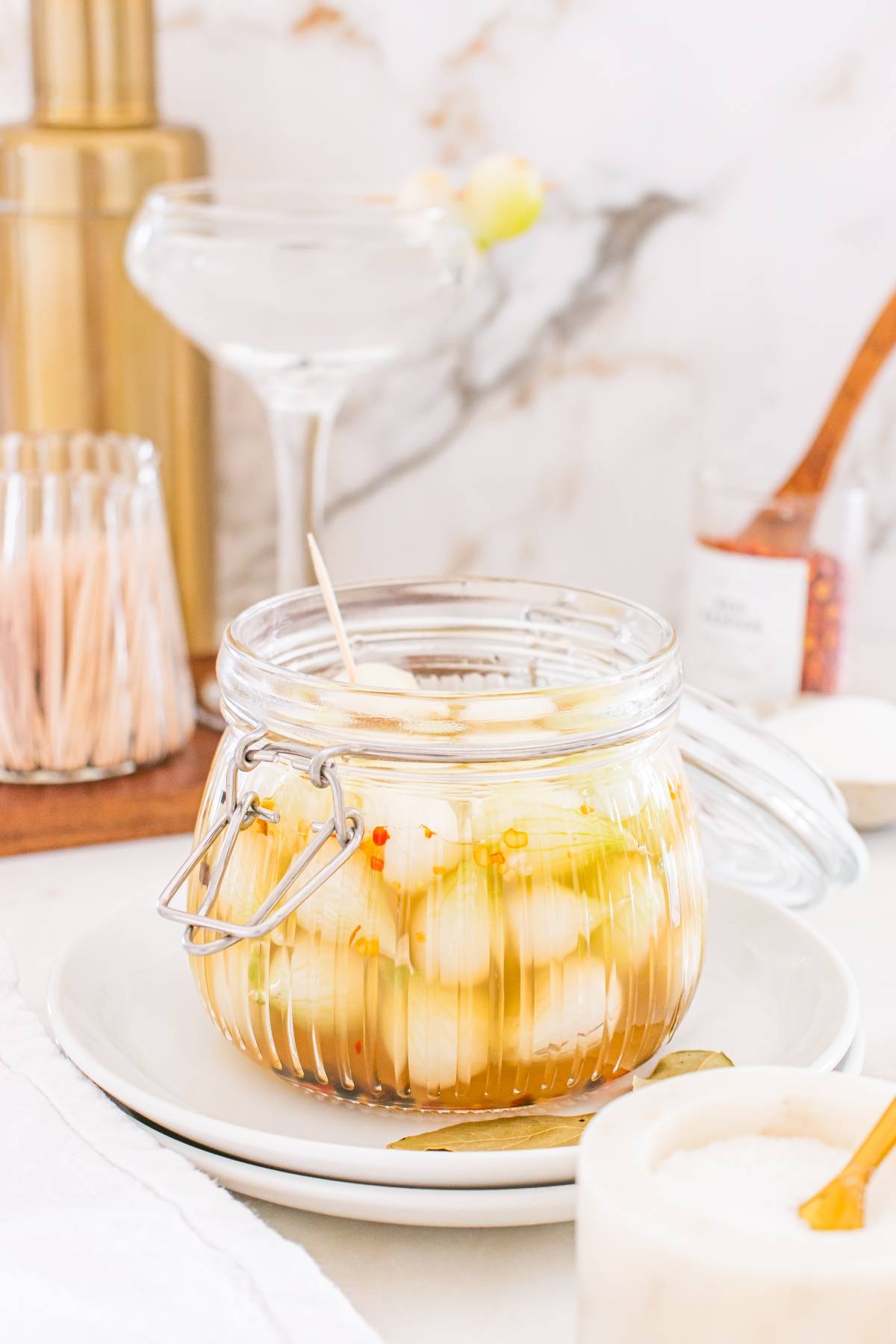 A glass jar of marinated cocktail onions with herbs and spices sits on a white plate with a toothpick.