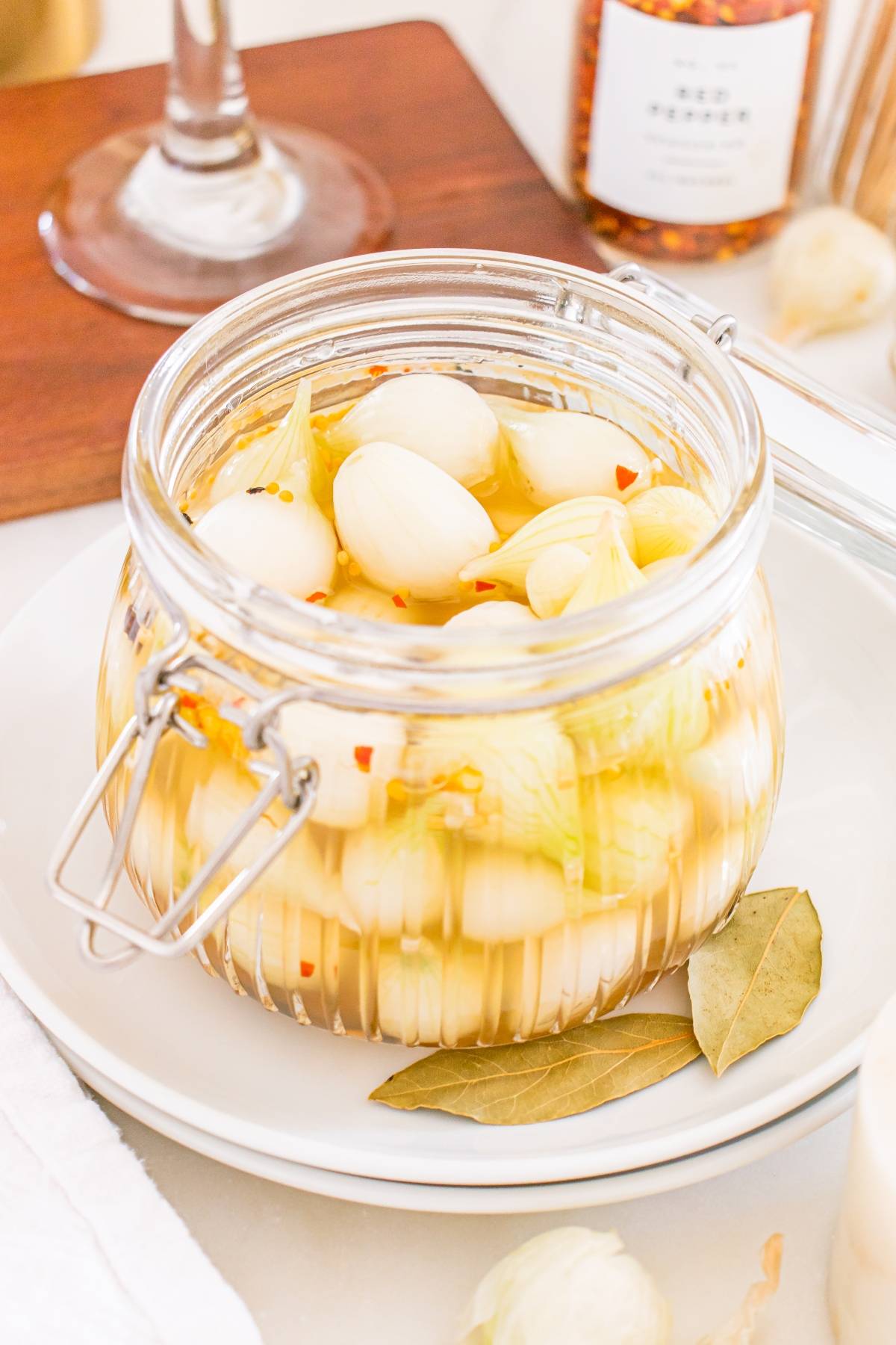 A jar of pickled onions with spices, bay leaves, and garlic on a white plate with a fork nearby.