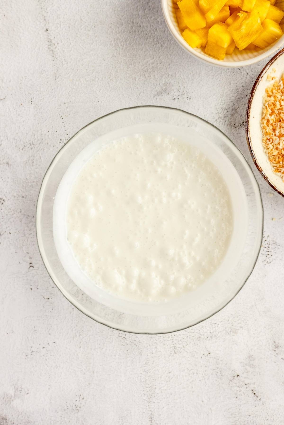 A glass bowl of creamy white batter sits beside bowls of pineapple and toasted coconut—perfect for making coconut pineapple frozen yogurt at home.