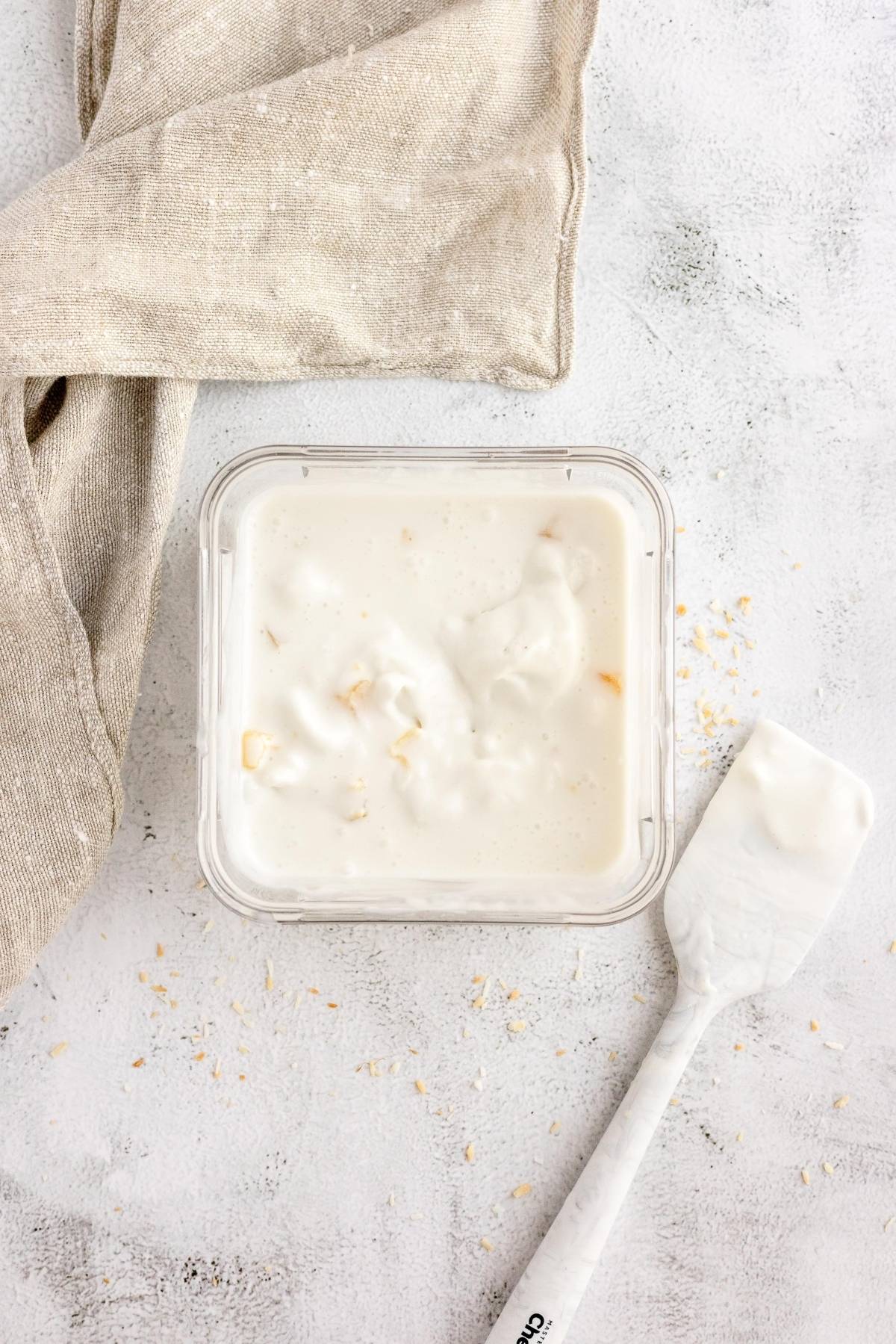 A container of creamy yogurt with a spatula and beige napkin on a light textured surface—perfect for anyone eager to make froyo or try the best frozen yogurt recipe at home.