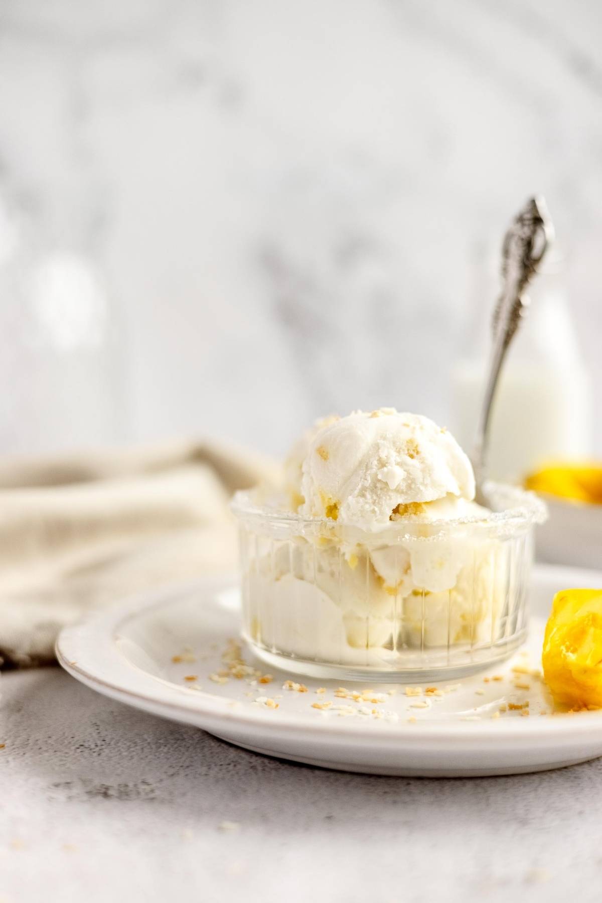 Two scoops of vanilla ice cream in a glass bowl on a white plate, with a spoon and mango pieces nearby—perfect inspiration to make froyo or enjoy as a Piña Colada frozen treat.