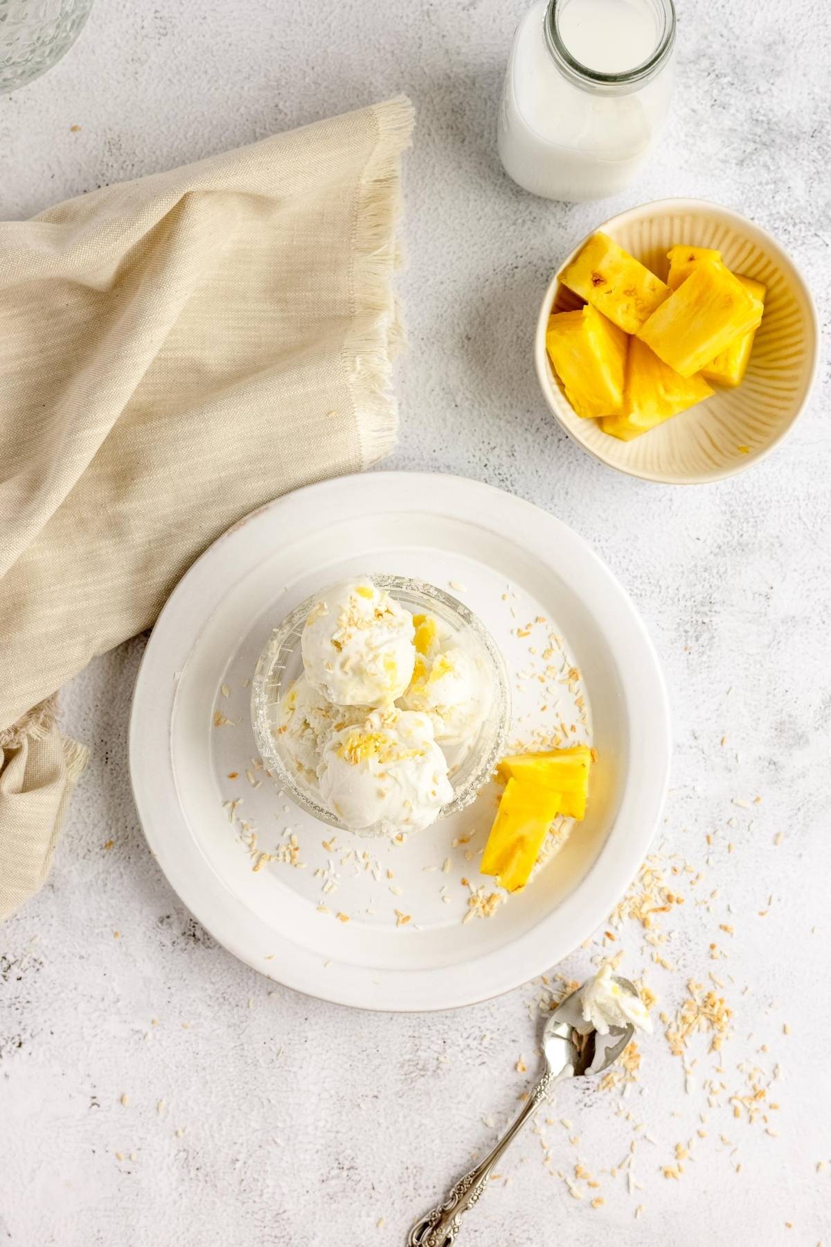 A bowl of vanilla frozen yogurt ice cream with pineapple chunks and shredded coconut on a white plate, next to milk and pineapple—perfect inspiration for the best frozen yogurt recipe or a tropical coconut pineapple frozen yogurt.