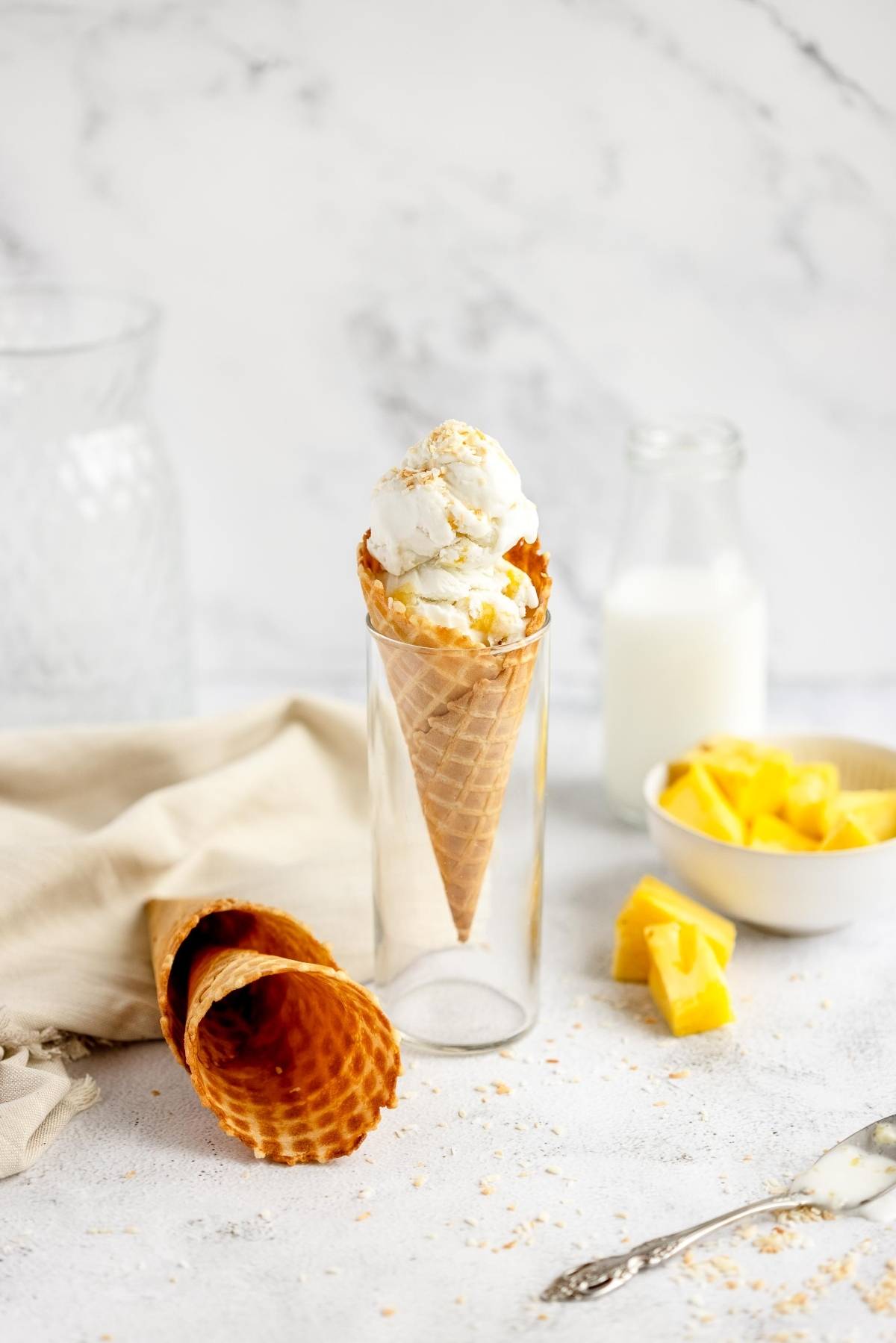 Vanilla ice cream in a waffle cone, blended with milk, pineapple chunks, and another cone on a light surface—a Piña Colada frozen treat that’s as refreshing as coconut pineapple frozen yogurt.