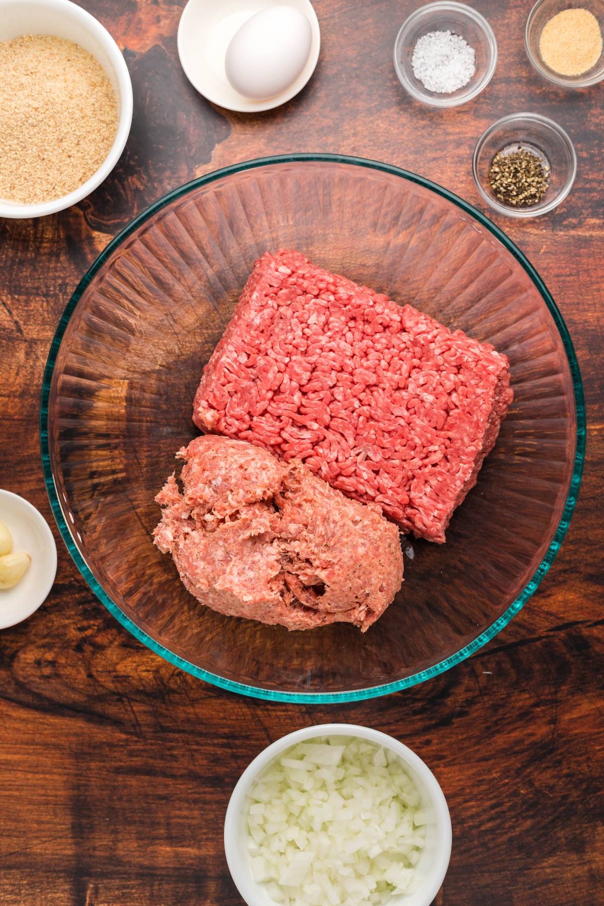 Ground beef and sausage in a glass bowl surrounded by breadcrumbs, egg, spices, garlic, and chopped onion—perfect for creating a delicious Swedish meatball recipe or your own copycat Ikea Swedish meatballs recipe.