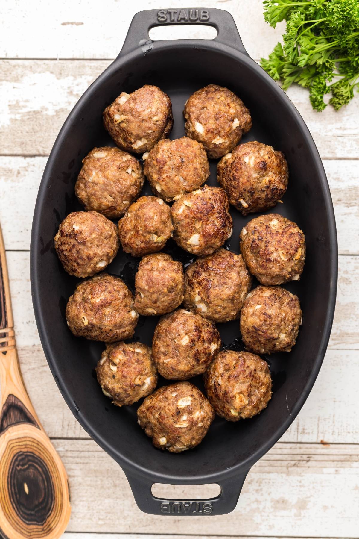 A black oval dish filled with cooked meatballs, next to a wooden spoon and fresh parsley—perfect for trying your favorite Swedish meatball recipe.